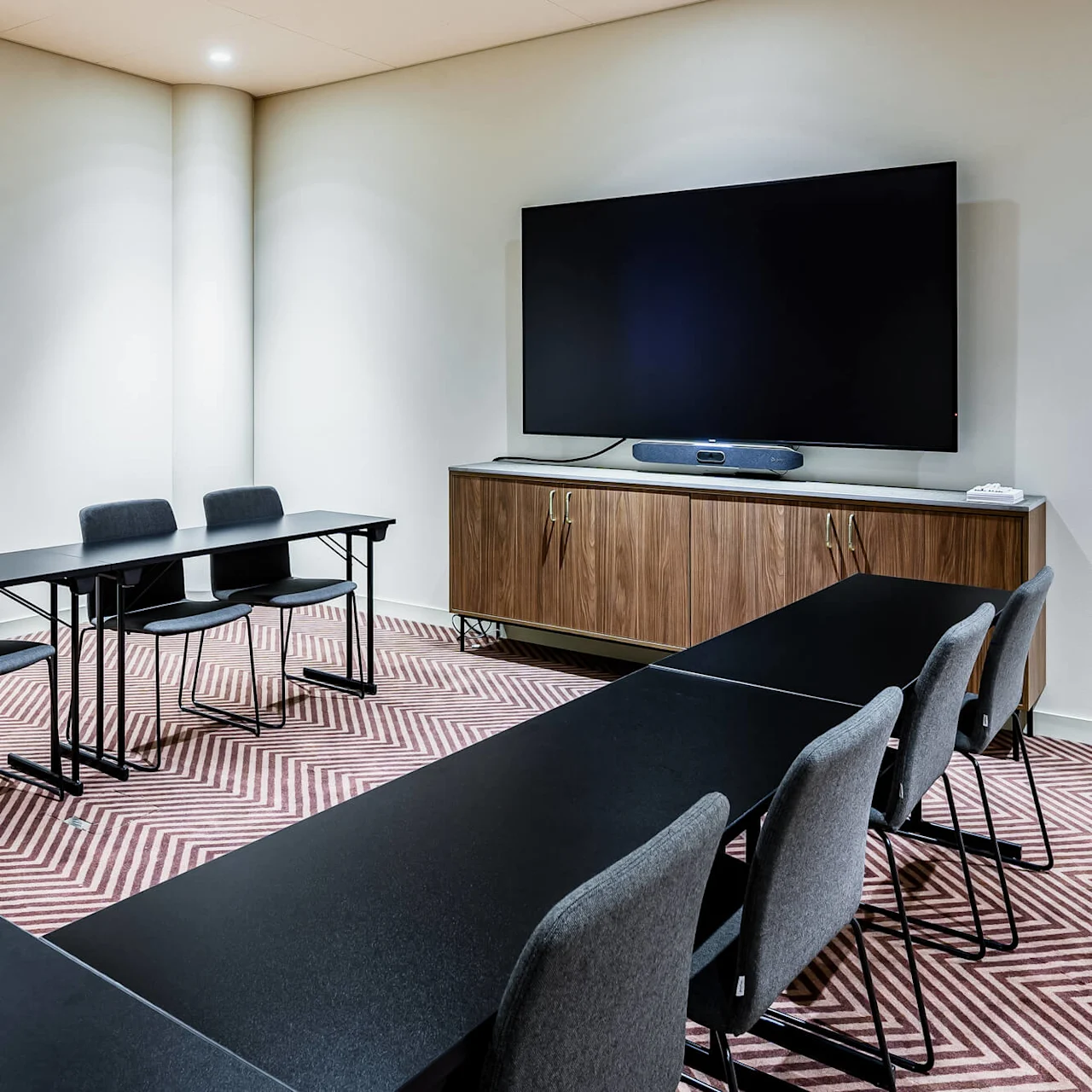 A conference room features a U-shaped table arrangement with black chairs. A large screen is mounted on the wall above a wooden cabinet. The floor has a striped pattern.