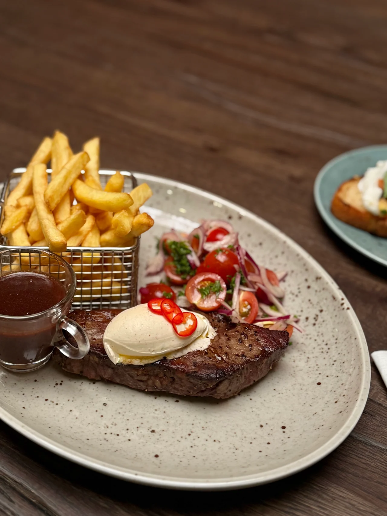 Steak topped with butter and chili peppers sits on a plate with a side salad and fries in a basket, accompanied by a small cup of sauce on a wooden table.