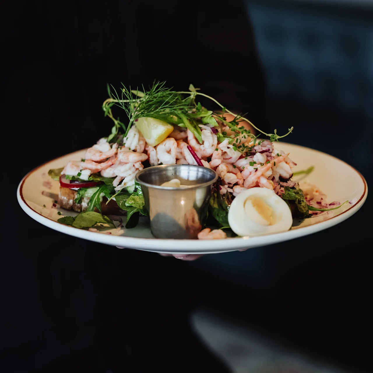 A plate of shrimp salad with lemon wedges, fresh greens, and a side of dressing is held by a person in a dimly lit restaurant.
