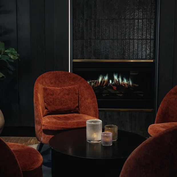 Et hyggeligt loungeområde med rustfarvede lænestole omkring et sofabord med stearinlys, mod en mørk væg med pejs.