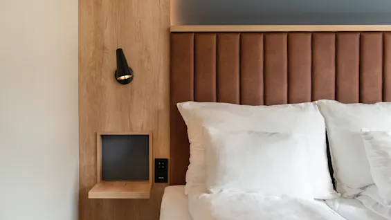 Et hyggeligt hotelværelse med en seng med brun sengegavl, hvidt sengetøj og et træpanel med en moderne sort lampe.
