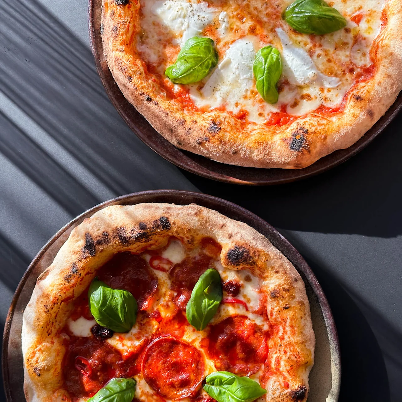 Two pizzas with crispy crusts, topped with melted cheese, tomato sauce, basil leaves, and pepperoni, rest on dark plates, illuminated by sunlight on a table.