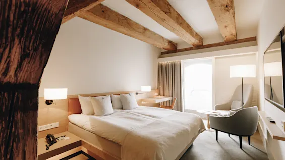 A cozy hotel room with a large bed, wooden ceiling beams, and a bright window. A desk, chairs, and a TV complete the comfortable space.