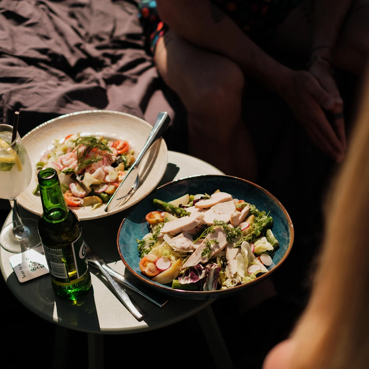 Två skålar med färsk sallad står på ett litet utomhusbord, tillsammans med en citroncocktail och en grön flaskdryck. Människor sitter i närheten i soligt väder.
