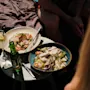 Två skålar med färsk sallad står på ett litet utomhusbord, tillsammans med en citroncocktail och en grön flaskdryck. Människor sitter i närheten i soligt väder.