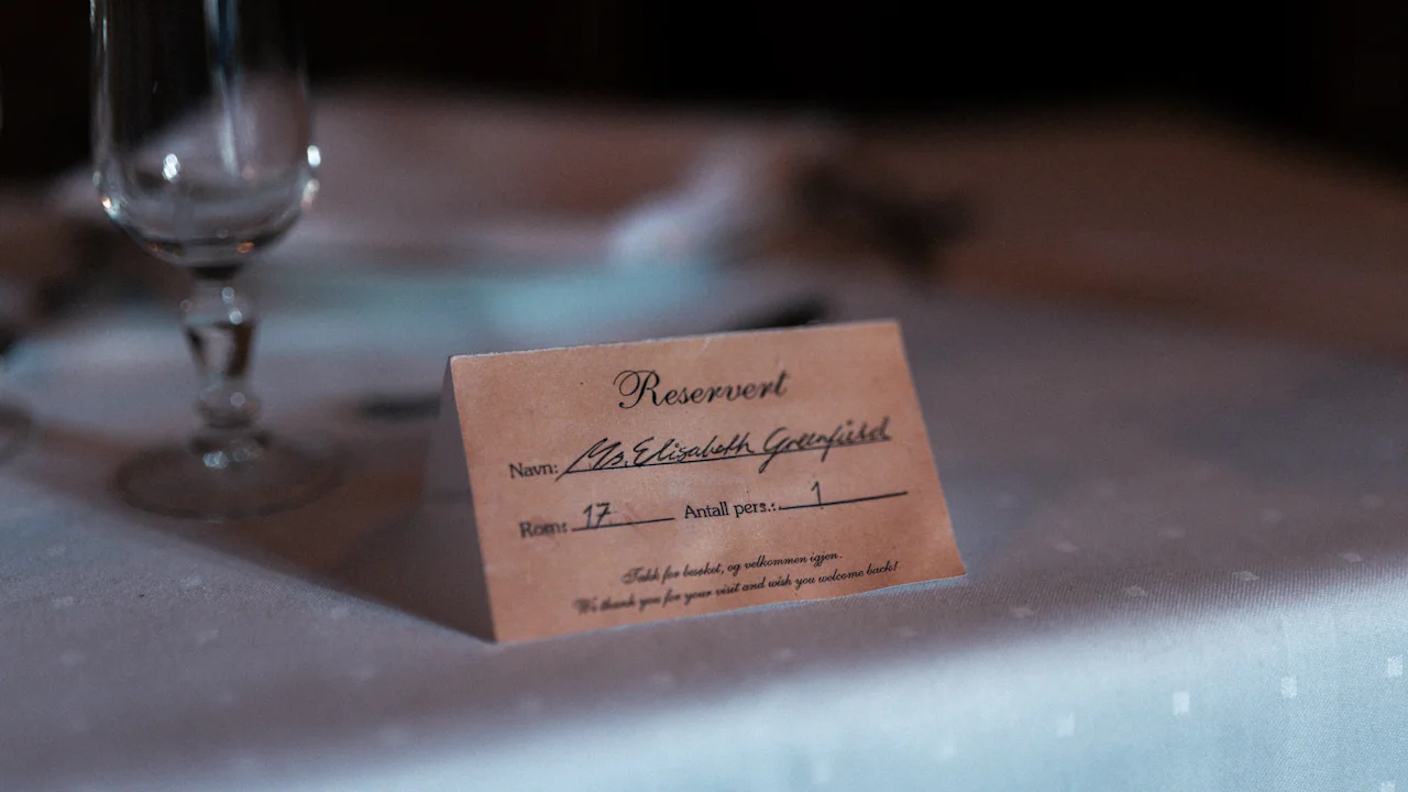 A reservation card at a dimly lit table in Dalen Hotel reads “Ms. Elizabeth Greenfield” with room and seating details, adding a mysterious touch to the elegant setting.