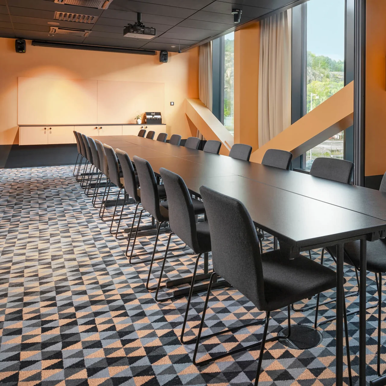 Ett långt konferensbord med svarta stolar runt det, beläget i ett mötesrum med geometrisk matta och stora fönster. Väggarna är beige med monterade högtalare och projektor.