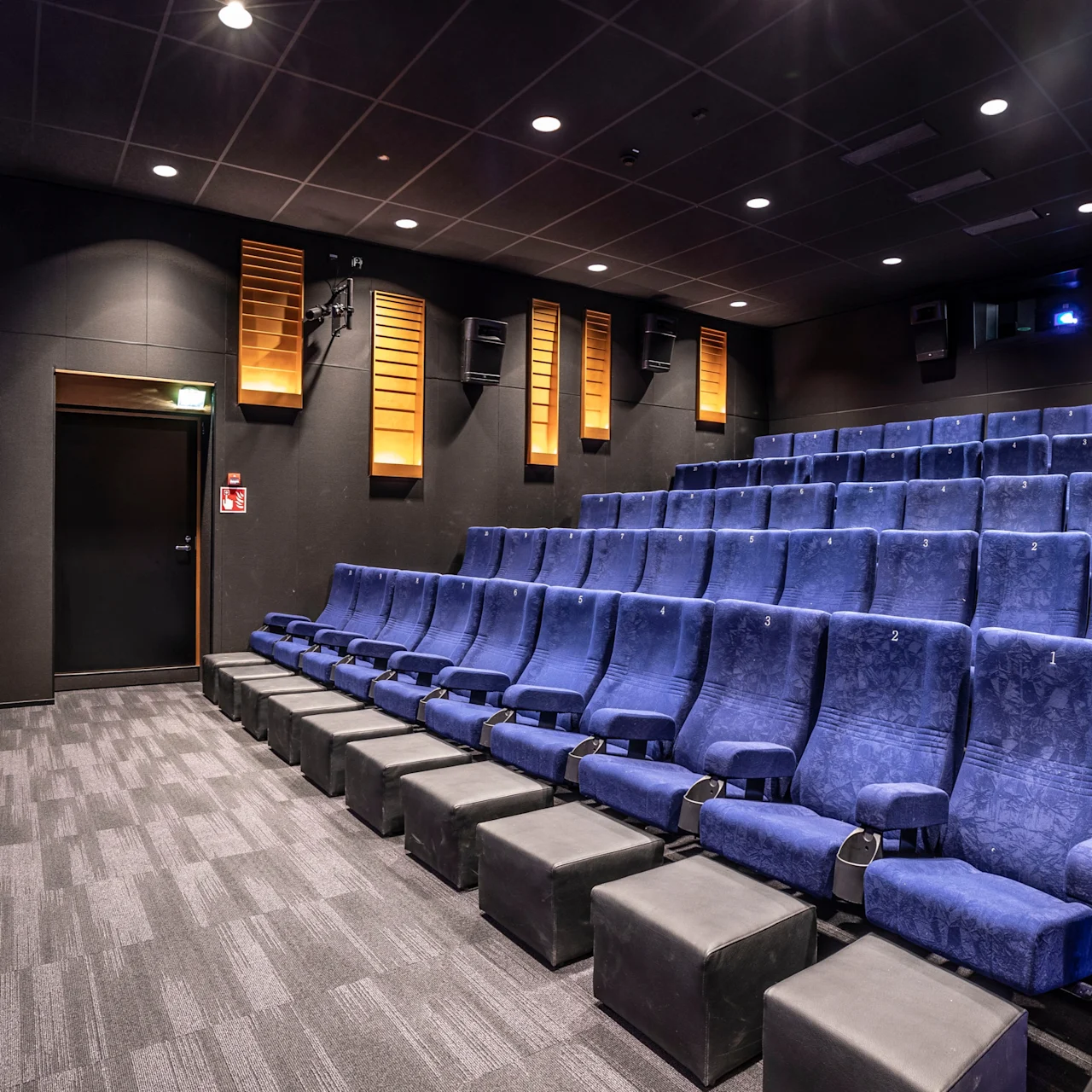 A small, empty cinema with tiered blue seats faces a large screen displaying "bobs." Walls are dark with orange acoustic panels, and stairs feature blue LED lights.