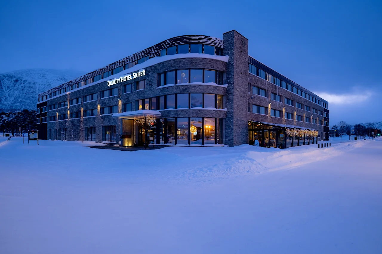 Et snødekket hotell med skiltet "QUALITY HOTEL SOGN" er opplyst mot en blå skumringshimmel, omgitt av snødekte fjell og trær.