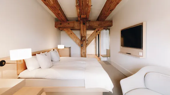 Ett modernt hotellrum med en stor säng, synliga träbjälkar i taket, en väggmonterad TV och en vit fåtölj.