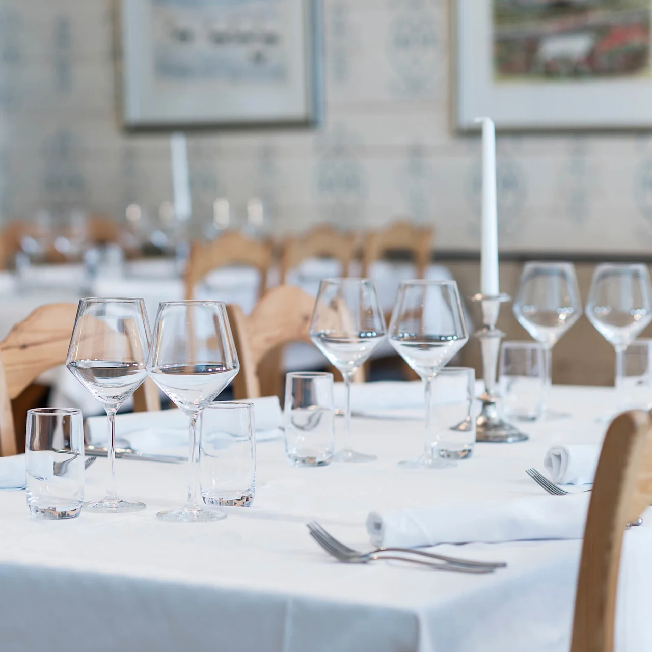 Ett bord elegant dukat med vinglas, ljus och vita servetter, omgivet av trästolar i en charmig restaurangmiljö med inramad konst på väggarna.
