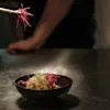En sort skål fylt med nudler toppet med spirer og rosa pynt; spisepinner holder mer pynt over. En person står uklar i bakgrunnen, plassert på en metallisk overflate.