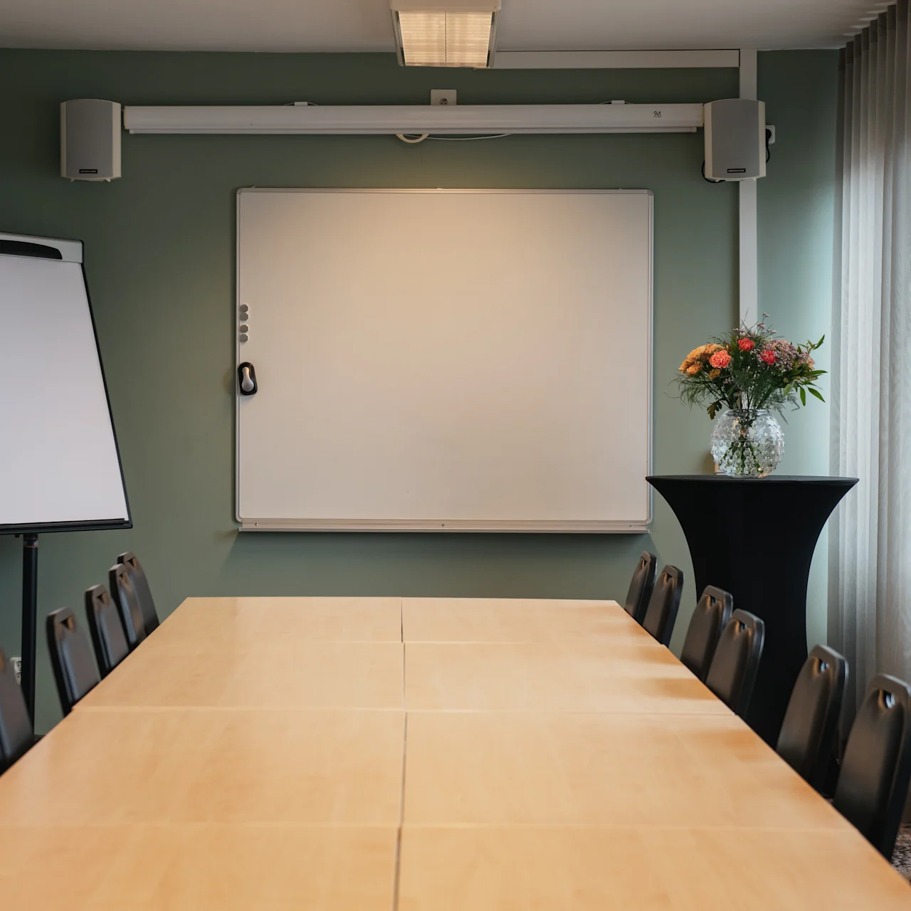 A long wooden conference table with chairs is in a room, facing a blank whiteboard. A flipchart and flowers on a tall table are nearby, with curtains along one wall.