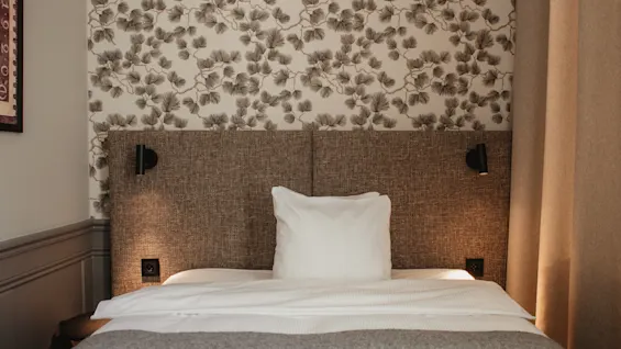 En bekväm hotellsäng med mönstrad tapet och varm belysning på Home Hotel Post i Oskarshamn.