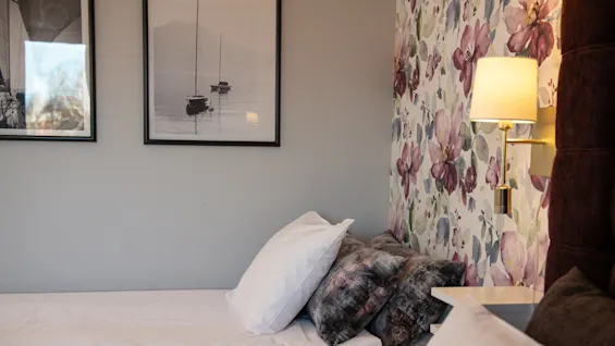 A neatly made bed with white and patterned cushions rests in a room with floral wallpaper and framed sailboat photos, illuminated by a wall-mounted lamp.