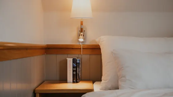 A cozy bedroom scene with a bedside table, books, and a warm lamp next to a bed with white linens.