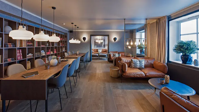 Spacious room with wooden tables and leather sofas arranged for meetings or relaxation. Bookshelves and hanging lamps provide a cozy, well-lit environment. Windows and plants add natural elements.