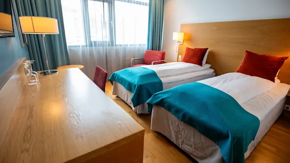 To pent oppredde enkeltsenger med turkise tepper og oransje puter står ved siden av et trebord med lampe og glass, i et hotellrom med store vinduer.