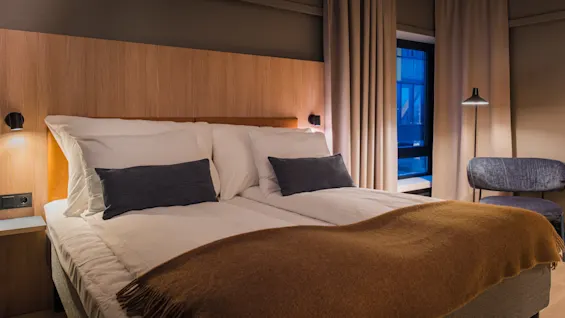 A cozy hotel room with a bed, bedside table, and a chair. The room is dimly lit, creating a relaxing atmosphere.