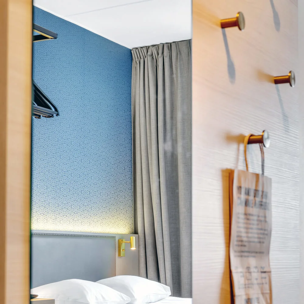 A neatly made bed with white linens is positioned against a blue textured wall, illuminated by a wall light. Curtains are drawn to one side. A paper bag hangs from a hook on the door.