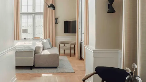 A modern living room features a beige sectional sofa, wall-mounted black lamp, and flat-screen TV, with large windows draped in peach curtains providing natural light. 