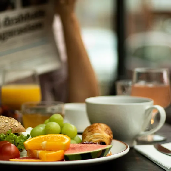 En tallrik med frukostmat, inklusive bröd, frukt och ett bakverk, står på ett bord. En person läser en tidning i bakgrunden. Text: "Så har vi drabbats av vardagsrasism".