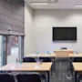 A conference room with a large screen on the wall, six desks arranged in rows, each with notepads, pens, and water bottles. Natural light comes through a window.