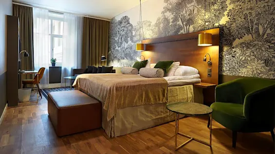 A well-lit bedroom featuring a neatly made bed with pillows, flanked by elegant nightstands and a forest-themed wallpaper accent wall.