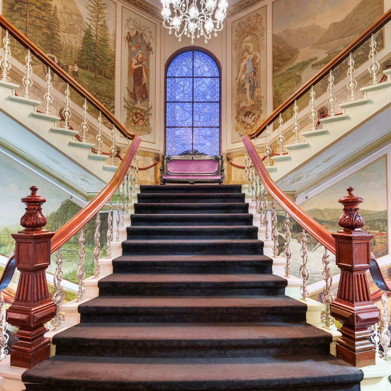 Suuri, matolla päällystetty portaikko, jossa on koristeelliset kaiteet, johtaa lasimaalaukseen ja seinämaalauksiin.