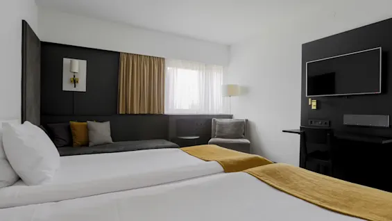A modern hotel room features two neatly made beds with mustard-colored accents, a wall-mounted TV, a seating area by the window, and a desk, creating a comfortable, sleek environment.
