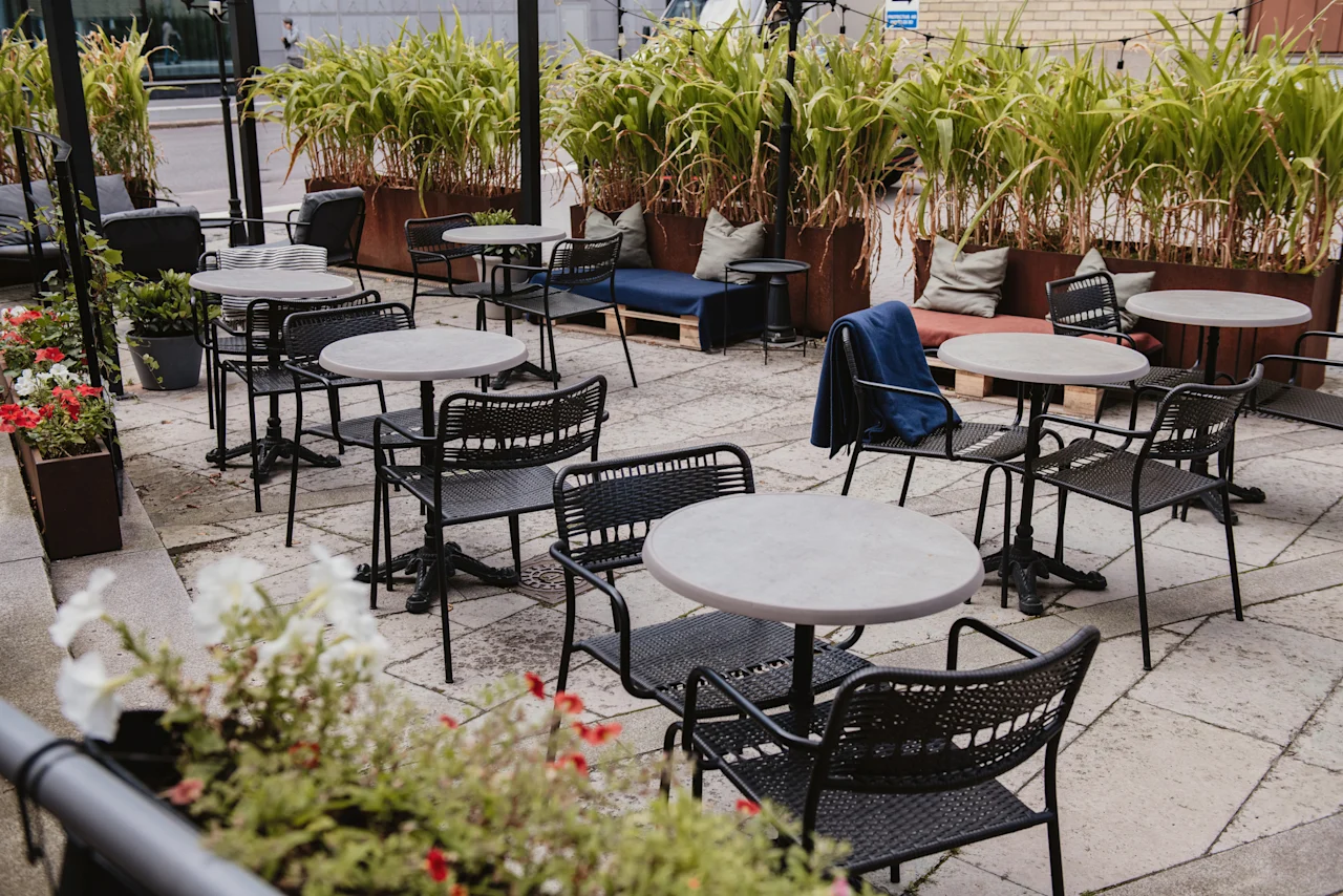 An inviting outdoor patio at Home Hotel Uppsala with several tables and chairs, surrounded by lush green plants and flowers.