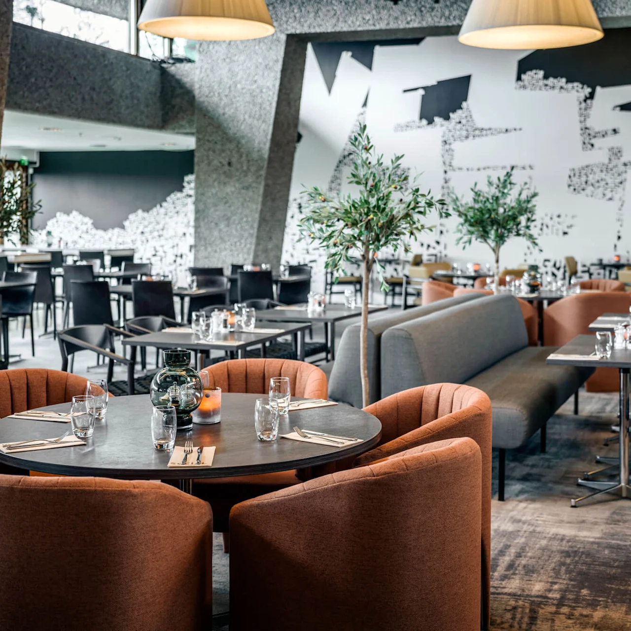 A round table with orange cushioned chairs is set for dining, surrounded by stylish modern decor, plants, and abstract wall art in an upscale restaurant environment.
