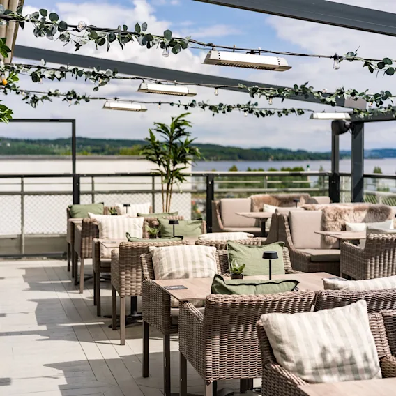Utendørs takterrasse med flettestoler og puter, pyntet med lyslenker og planter, samt utsikt over en innsjø.