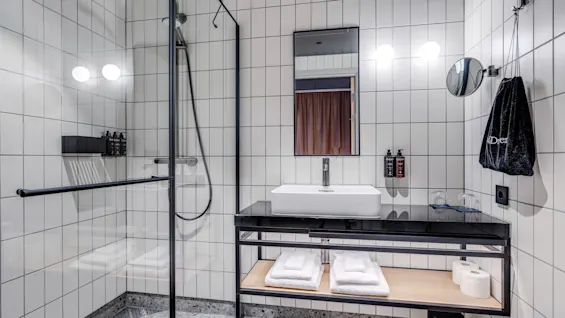 Bruser med regnhoved omgivet af glas; elegant moderne håndvask med spejl over; hvide håndklæder, pumpeflasker, sort taske på flisevægge.