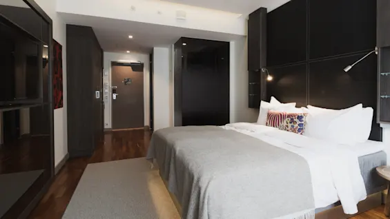 En pænt redt seng med hvide og farverige puder står i et moderne hotelværelse. Mørke træmøbler og blød belysning skaber en varm atmosfære.