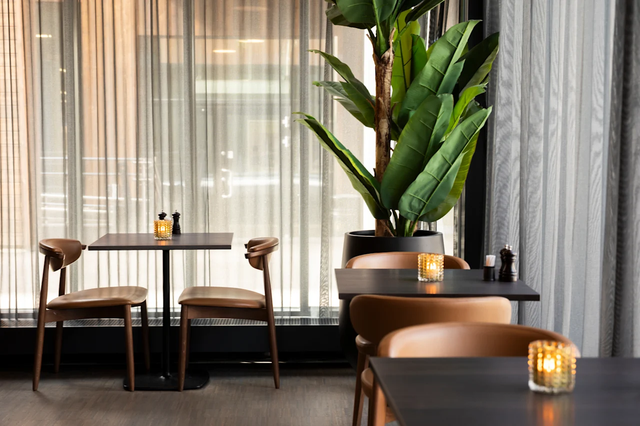 Et koselig restaurantinteriør med mørke bord, brune stoler og en stor grønn plante. Myk belysning fra stearinlys skaper en varm atmosfære.