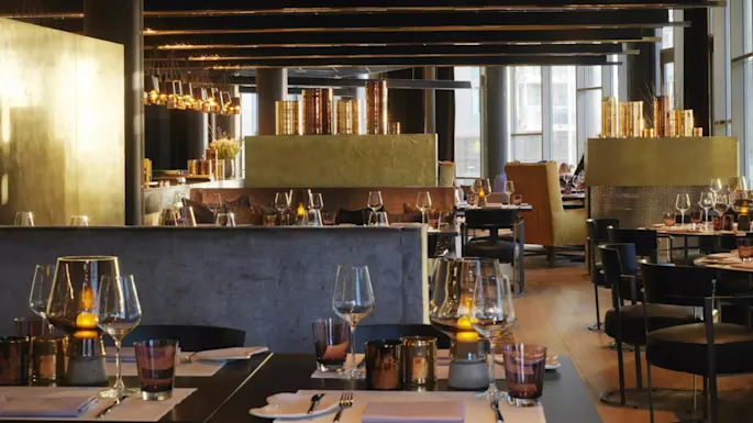 Restaurant tables arranged elegantly with wine glasses and candle holders, under modern lighting fixtures in a sophisticated, dimly lit urban setting. Large windows provide natural light in the background.