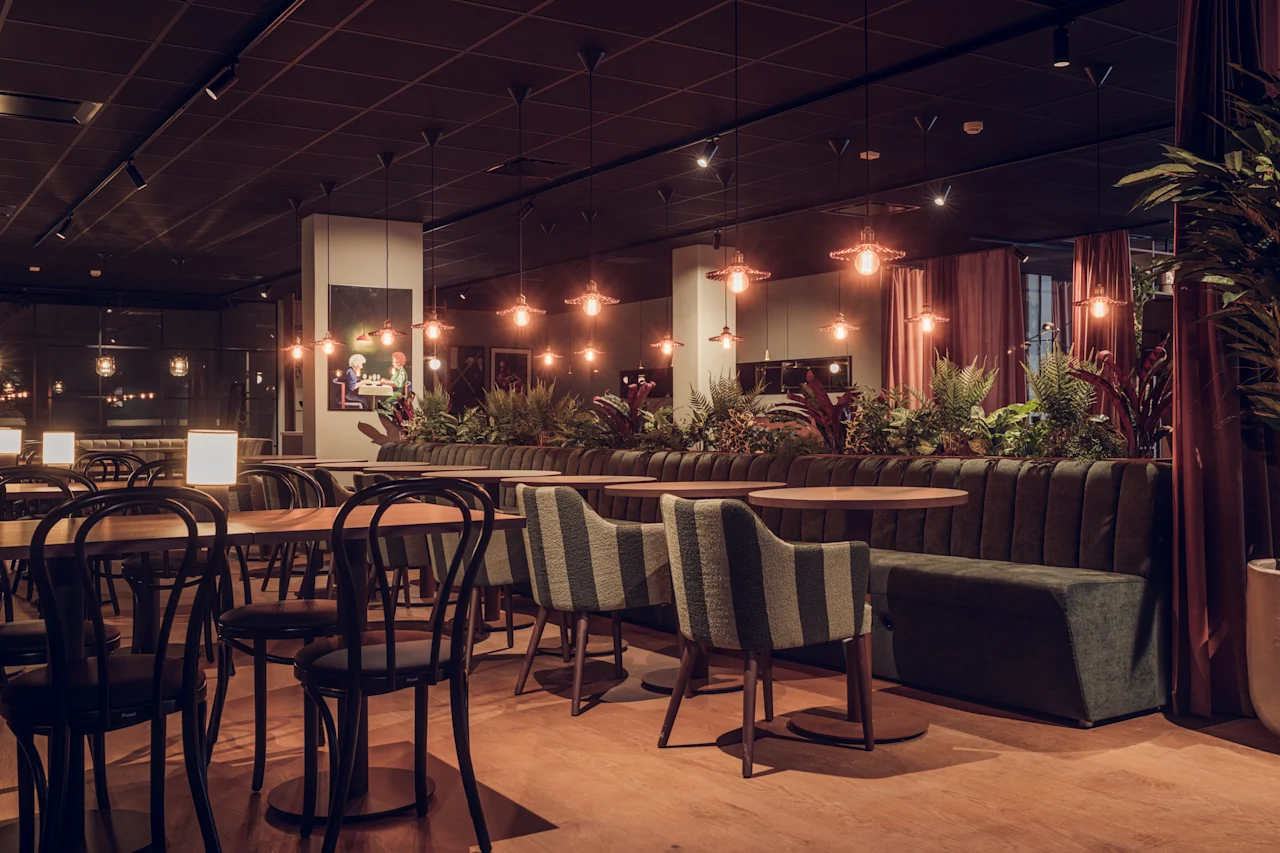 A restaurant features wooden tables and chairs arranged neatly, surrounded by soft lighting. Indoor plants adorn the space, creating a cozy ambiance with warm tones and modern decor.