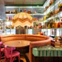 En buet bod med et rundt bord og farverige stole omgivet af eklektisk indretning og forskellige genstande på hylder, der skaber en livlig, hyggelig caféatmosfære.