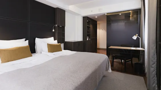 Seng med hvidt sengetøj og brune puder i et moderne hotelværelse. Mørke træmøbler, et skrivebord med lampe og blød belysning skaber en hyggelig atmosfære.
