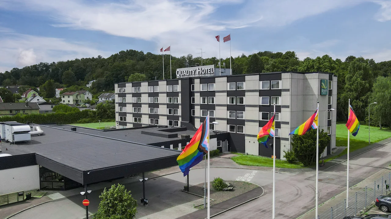 Ett fler våningshotell har regnbågsflaggor vid ingången, omgiven av träd och kullar. Text: "Quality Hotel."