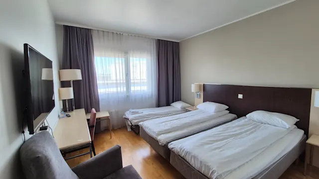 Hotellihuone, jossa kaksi erillistä vuodetta rivissä, työpöytä ja tuoli ison ikkunan edessä. TV on kiinnitetty seinälle, ja lähellä on nojatuoli.