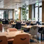 Moderni ravintolaruokailutila, jossa on paljon pöytiä ja tuoleja, kasveja, kynttilöitä ja suuret ikkunat. Poistumiskyltti on näkyvissä.