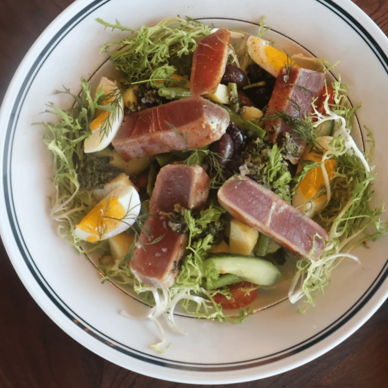 A salad with seared tuna, leafy greens, and sliced eggs, arranged on a white plate beside cutlery wrapped in a green holder on a wooden table. Initials "NDR" on holder.