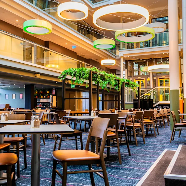 Ett rymligt hotellatrium med matbord, moderna cirkulära lampor och frodiga gröna växter, vilket skapar en inbjudande atmosfär.