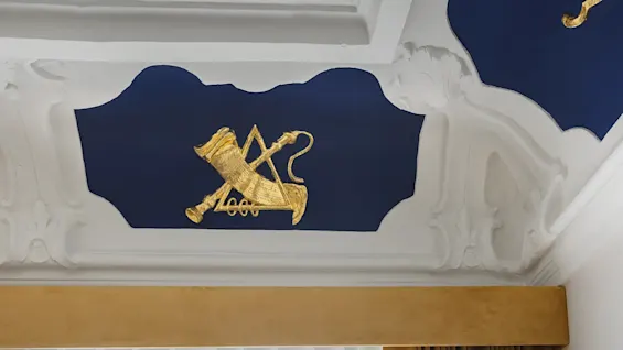 Et udsmykket loft med et gyldent emblem på et dybblåt panel, omgivet af hvid dekorativ stuk, over brune gardiner.