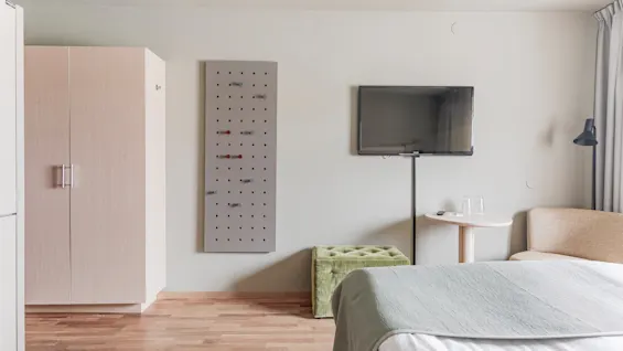 A modern hotel room with a bed, a wall-mounted TV, a wardrobe, and a pegboard for hanging items.