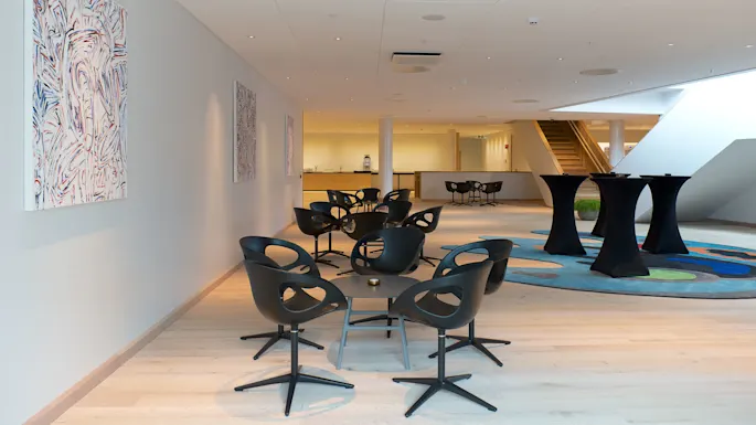 Modern black chairs surrounding small round tables in a spacious interior, featuring abstract paintings on white walls, high ceilings with recessed lighting, and a wooden staircase in the background.