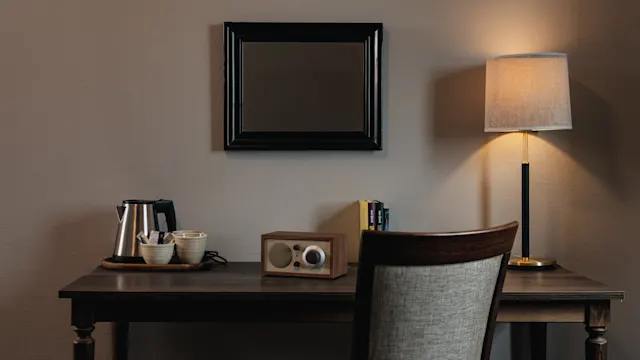 A wooden desk holds a kettle, two cups, a radio, and books. A lamp emits soft light beside a simple chair. Framed mirror hangs on the wall.