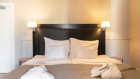 En bekväm hotellsäng med vita kuddar, en grå filt och vita tofflor, vilket antyder en avkopplande vistelse.